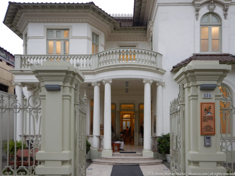 White historic residence behind ornamental gate with paired columns, curved balustraded balcony, and recessed entrance.