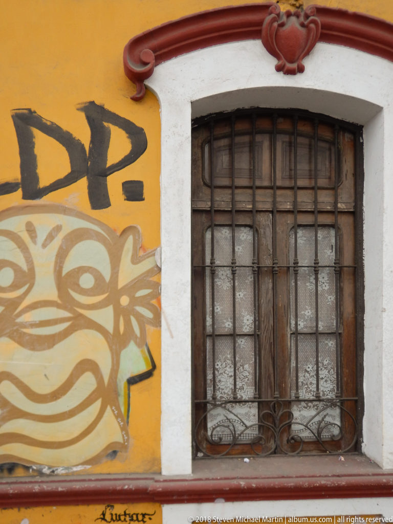 Weathered barred wooden window with white-and-red molded surround beside black and tan graffiti on an ochre wall.