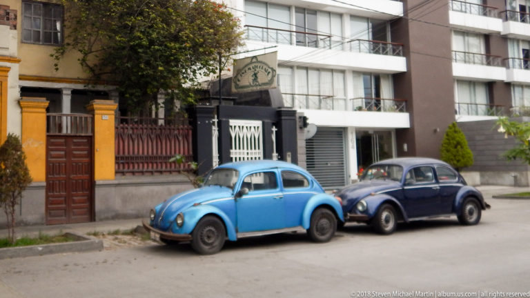 Bright blue and dark blue classic Volkswagen Beetles parked nose-to-tail beside mixed residential buildings.