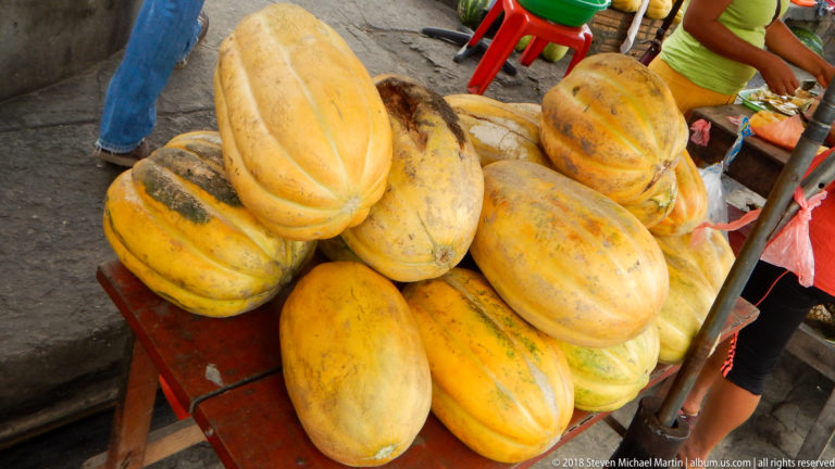 Large yellow papayas with green mottling are stacked on a low red market table.