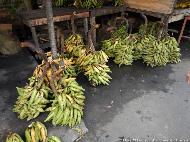 Large bunches of green bananas or plantains lie on the pavement beneath a wooden market table.