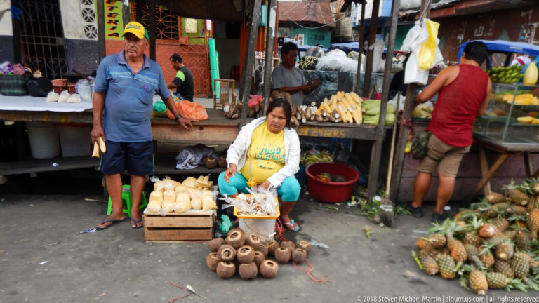 Seated market vendor sits beside brown root crops, a crate of pale produce, and a pile of pineapples.