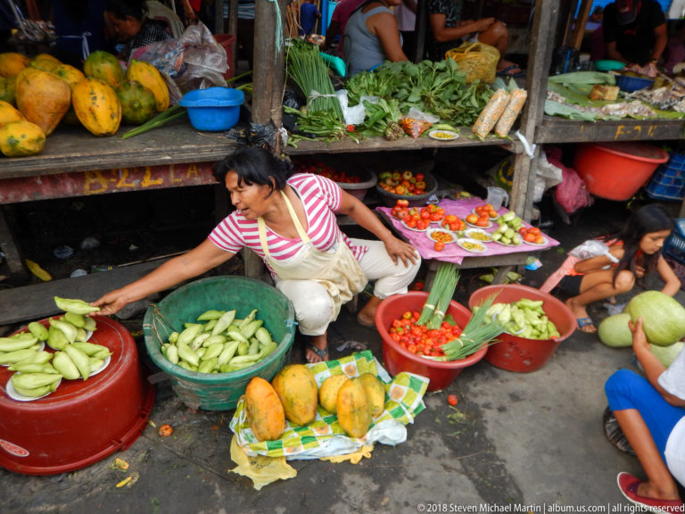 Woman vendor reaches across tubs of green vegetables and tomatoes beside papayas at a crowded produce stall.