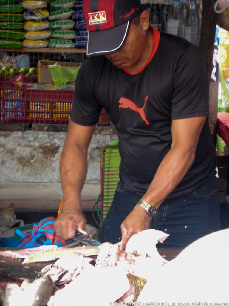 Market worker cuts a large fish with a knife on a busy preparation table.