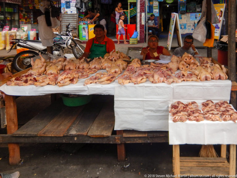 Raw poultry carcasses and cut pieces cover white-topped tables while three vendors sit behind the stall.