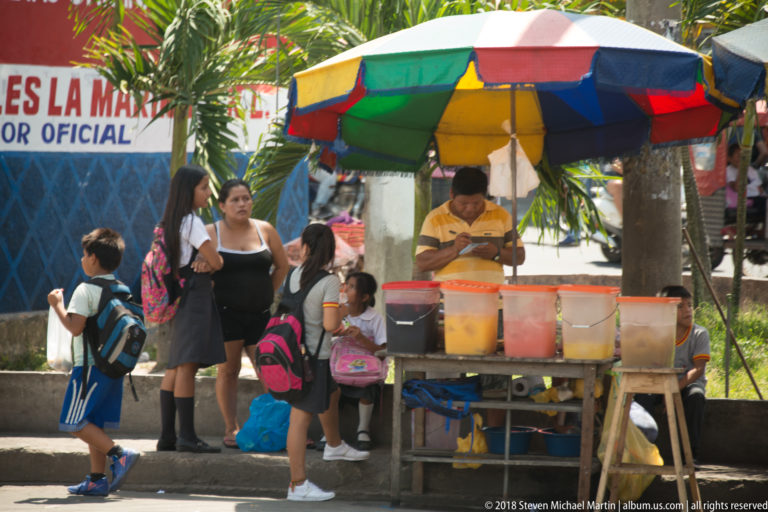 Vendor stands behind containers of colorful drinks beneath a multicolored umbrella as children and adults gather nearby.