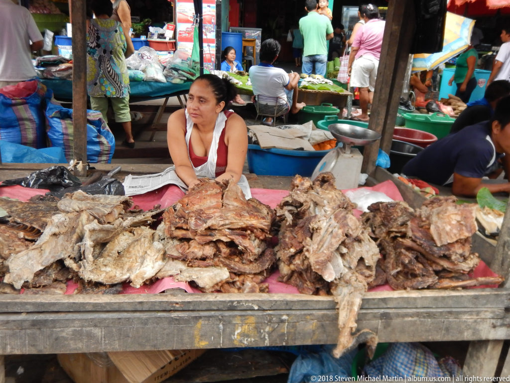 Seated woman vendor sits behind large piles of dried fish at a crowded indoor-outdoor market stall.