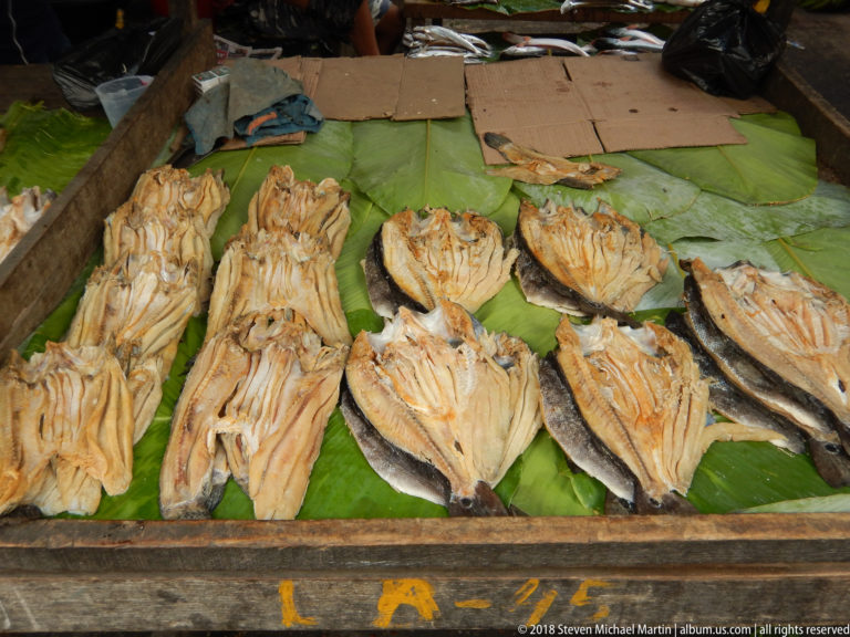 Split dried fish lie opened flat on broad green leaves across a wooden market counter.