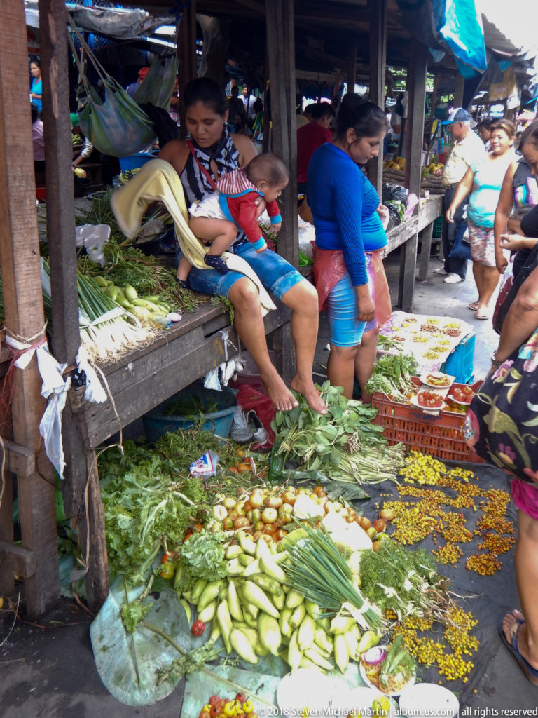 Vegetables and herbs cover broad leaves on the ground while vendors sit at a wooden stall and shoppers pass nearby.