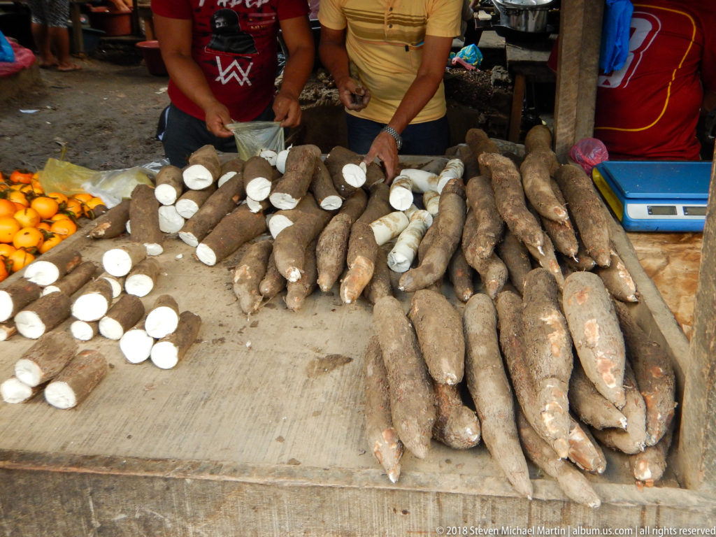 Whole brown cassava roots and cut white sections are piled across a wooden market table.