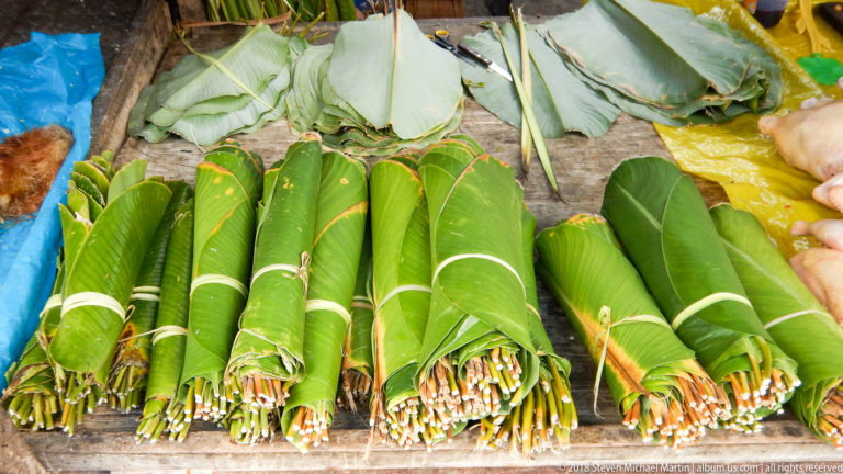 Rows of tightly tied green leaf bundles lie on a market table, with larger wrapping leaves stacked behind.