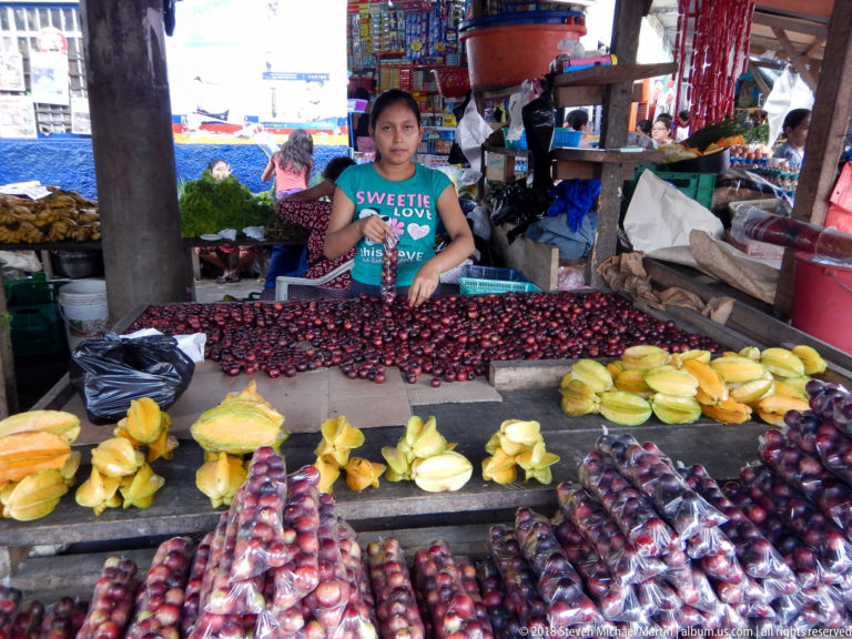 Vendor stands behind rows of yellow starfruit and dark red-purple tropical fruit at a crowded market stall.