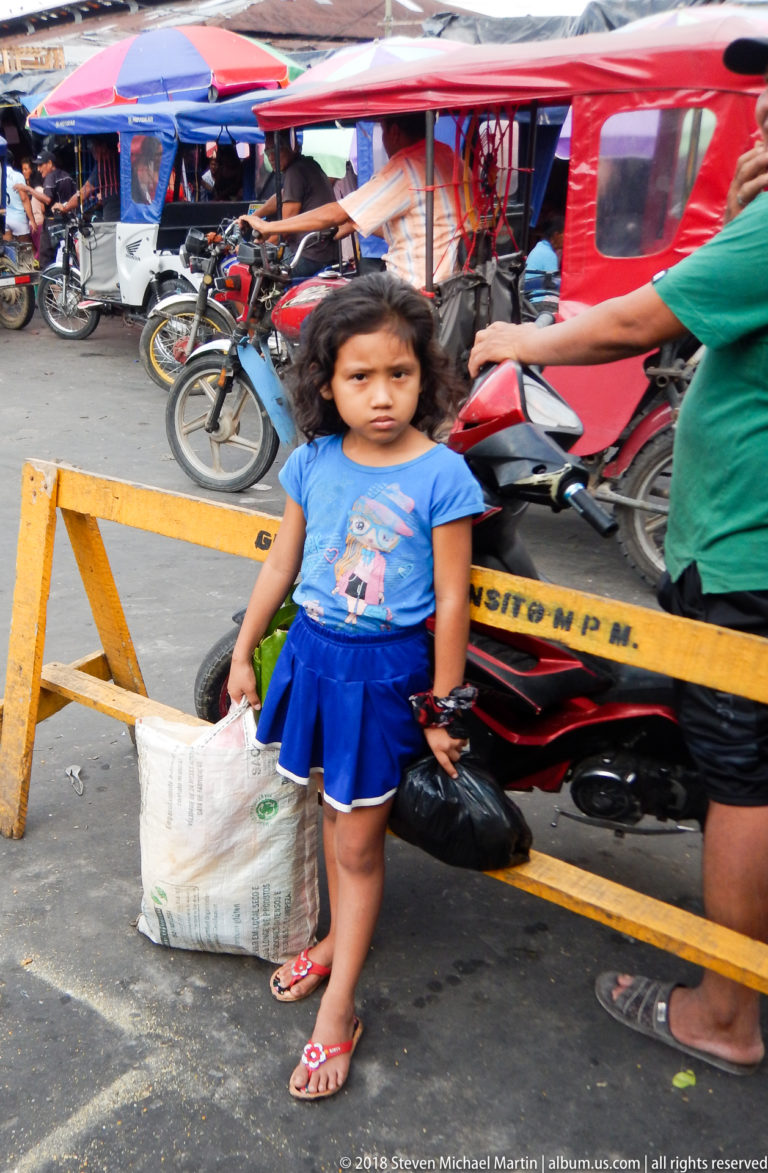 Young girl stands beside a yellow market barrier with bags while red mototaxis and motorcycles pass behind.