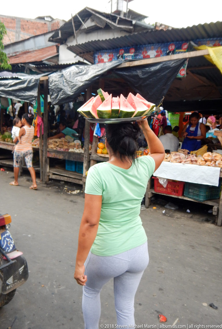 Woman in a green shirt carries a tray of watermelon slices balanced on her head through the market.