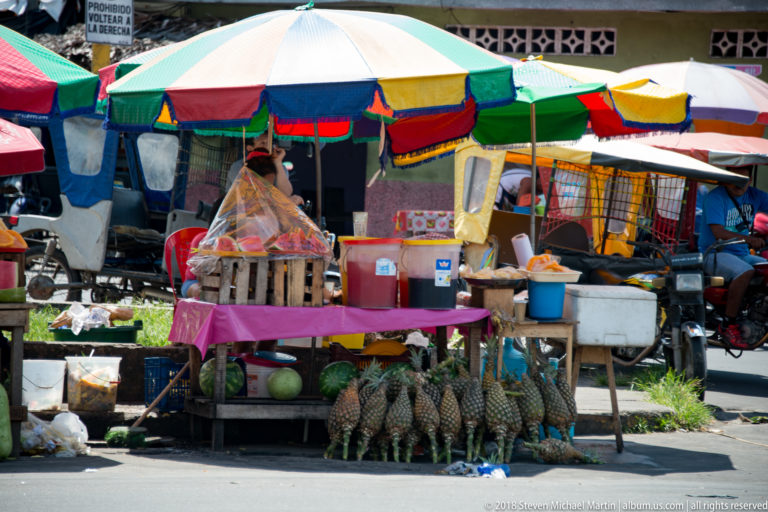 Roadside stall under a multicolored umbrella displays pineapples, green watermelons, containers, and hanging goods.