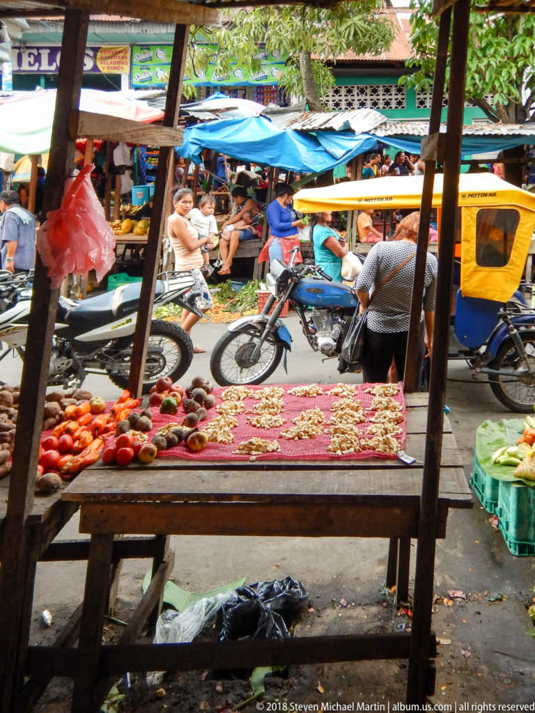 Low market table of red produce stands beside a street crowded with motorcycles, mototaxis, vendors, and blue tarps.