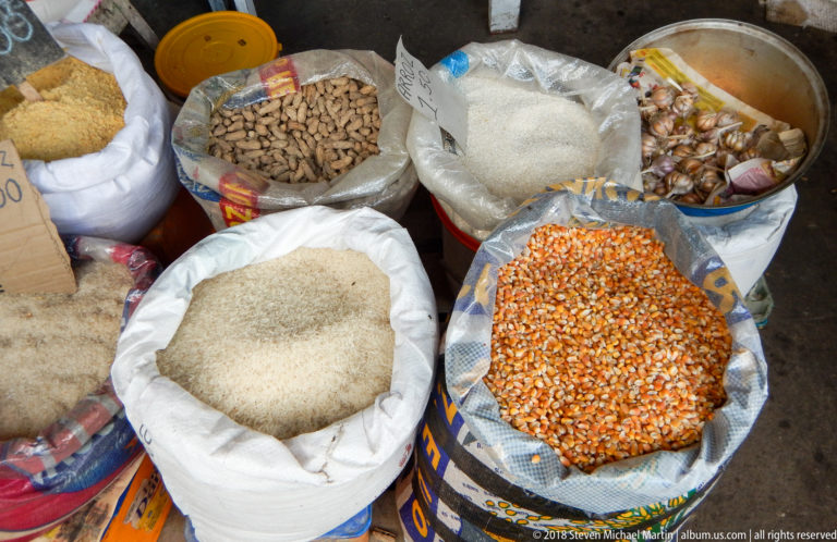 Open sacks hold white rice, orange corn kernels, peanuts, and other dry staples at a market stall.