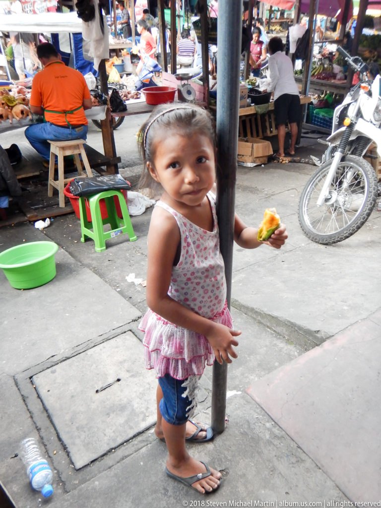 Young girl stands beside a metal post eating, with produce stalls, stools, and a motorcycle behind her.