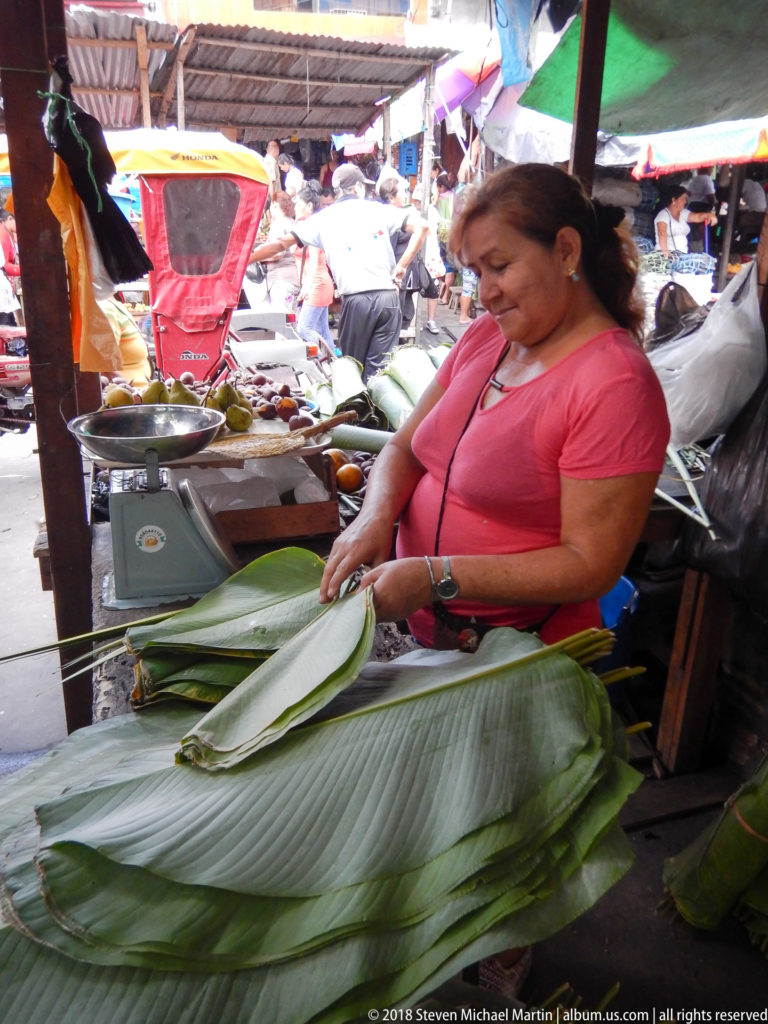 Vendor trims and stacks broad green leaves beside a tabletop scale at an Iquitos Market stall.