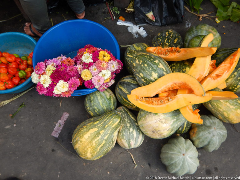 Orange squash wedges rest on whole striped gourds beside tubs of tomatoes and bright market flowers.
