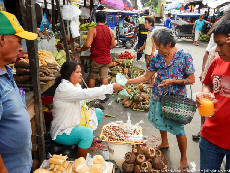 Seated vendor hands produce to an older woman with a basket amid tomatoes, pineapples, roots, and other market foods.