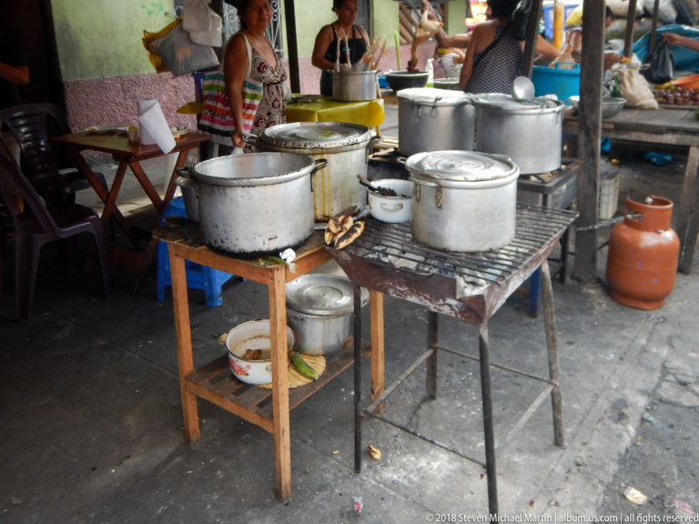 Large aluminum cooking pots stand on improvised tables beside a gas cylinder inside a crowded market food stall.