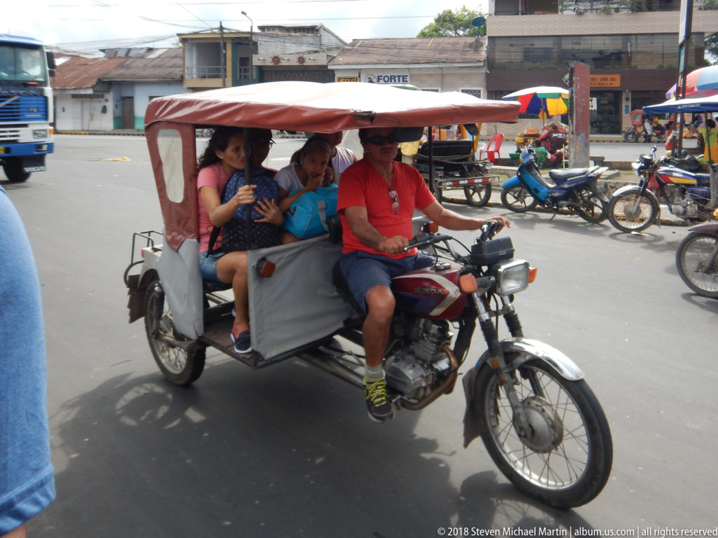 Red-and-white three-wheeled motorcycle taxi carries passengers past motorcycles and market stalls on a busy Iquitos street.