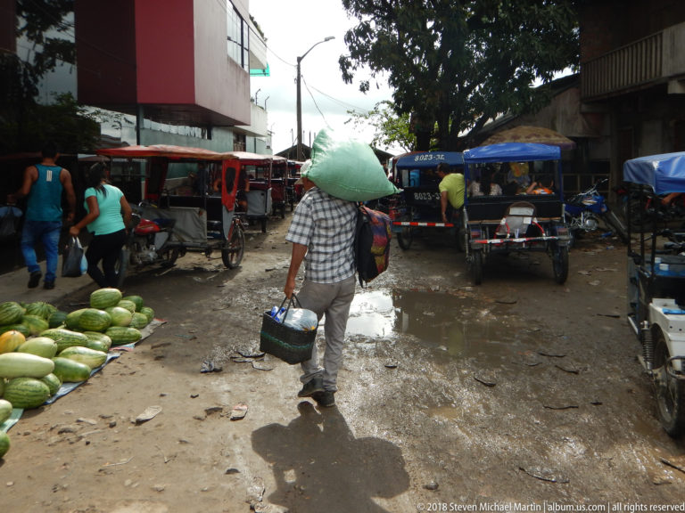 Shopper in a conical hat walks through a wet market lane beside green produce and parked three-wheeled mototaxis.