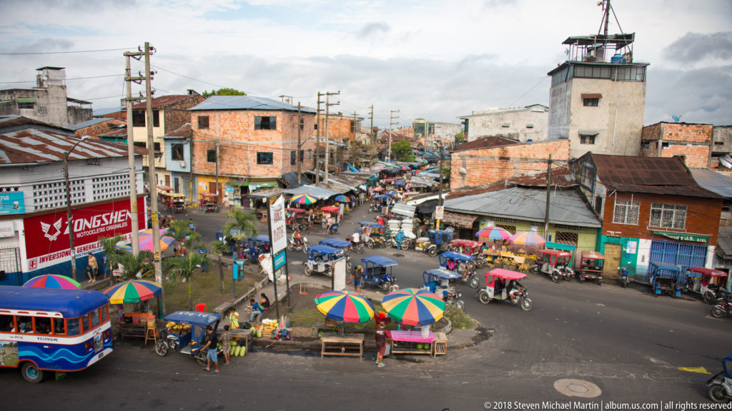 Elevated view of a market intersection with colorful umbrellas, mototaxis, low brick buildings, and a tall utilitarian tower.