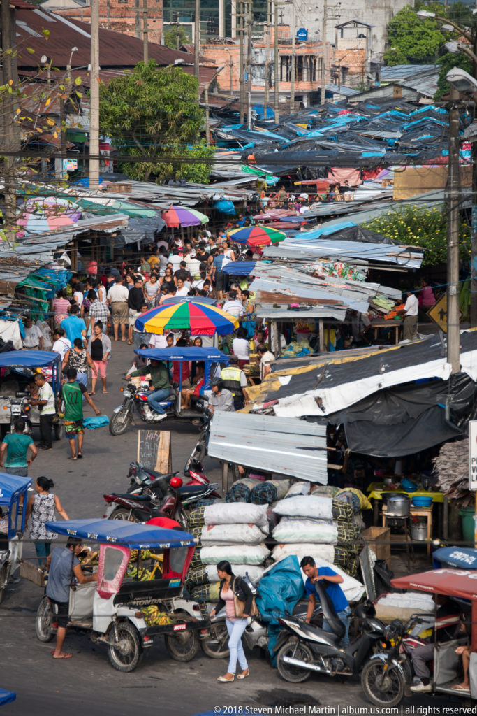 Elevated view down a crowded Iquitos market street filled with pedestrians, mototaxis, motorcycles, corrugated stalls, and colorful umbrellas.