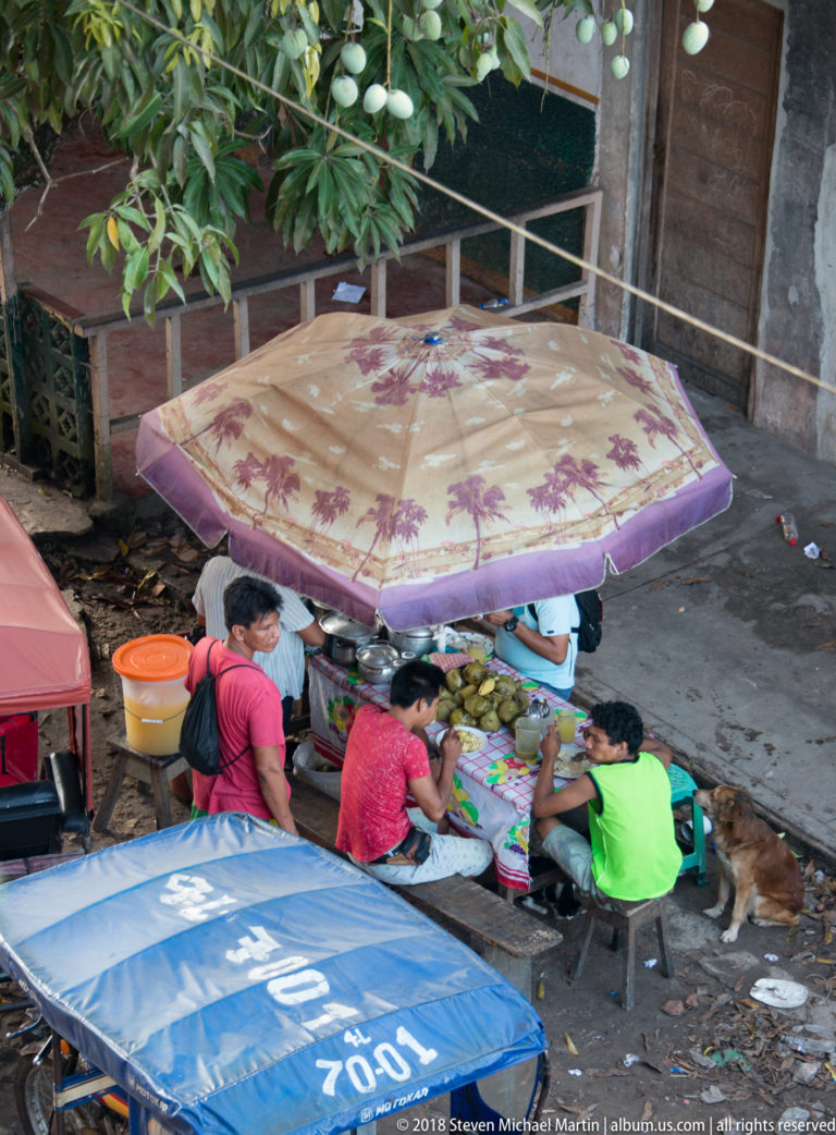 Several people sit around a small produce stall beneath a large patterned umbrella, with buckets, a motorcycle, and neighboring canopies nearby.