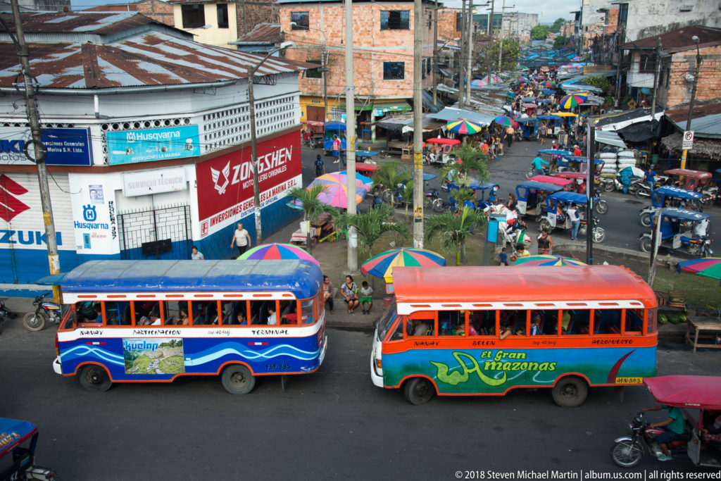 Two colorful buses pass below an elevated viewpoint beside a dense market of umbrellas, motorcycles, pedestrians, and brick buildings.