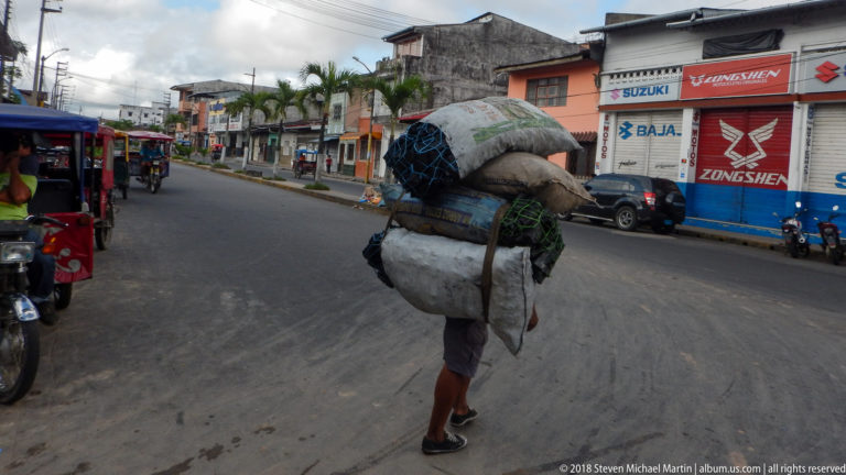 Market porter bends beneath stacked sacks and bundled vegetation while crossing a street lined with mototaxis and motorcycle shops.