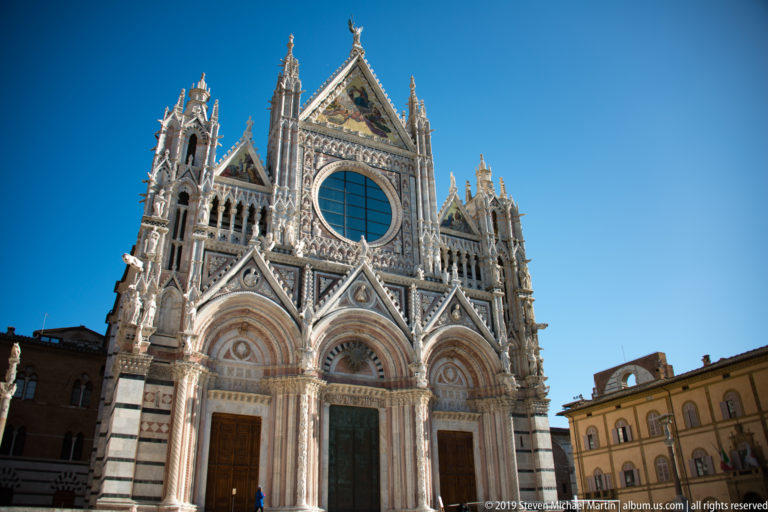 Siena Cathedral’s west façade rises beside neighboring buildings, with a visitor at its portals.