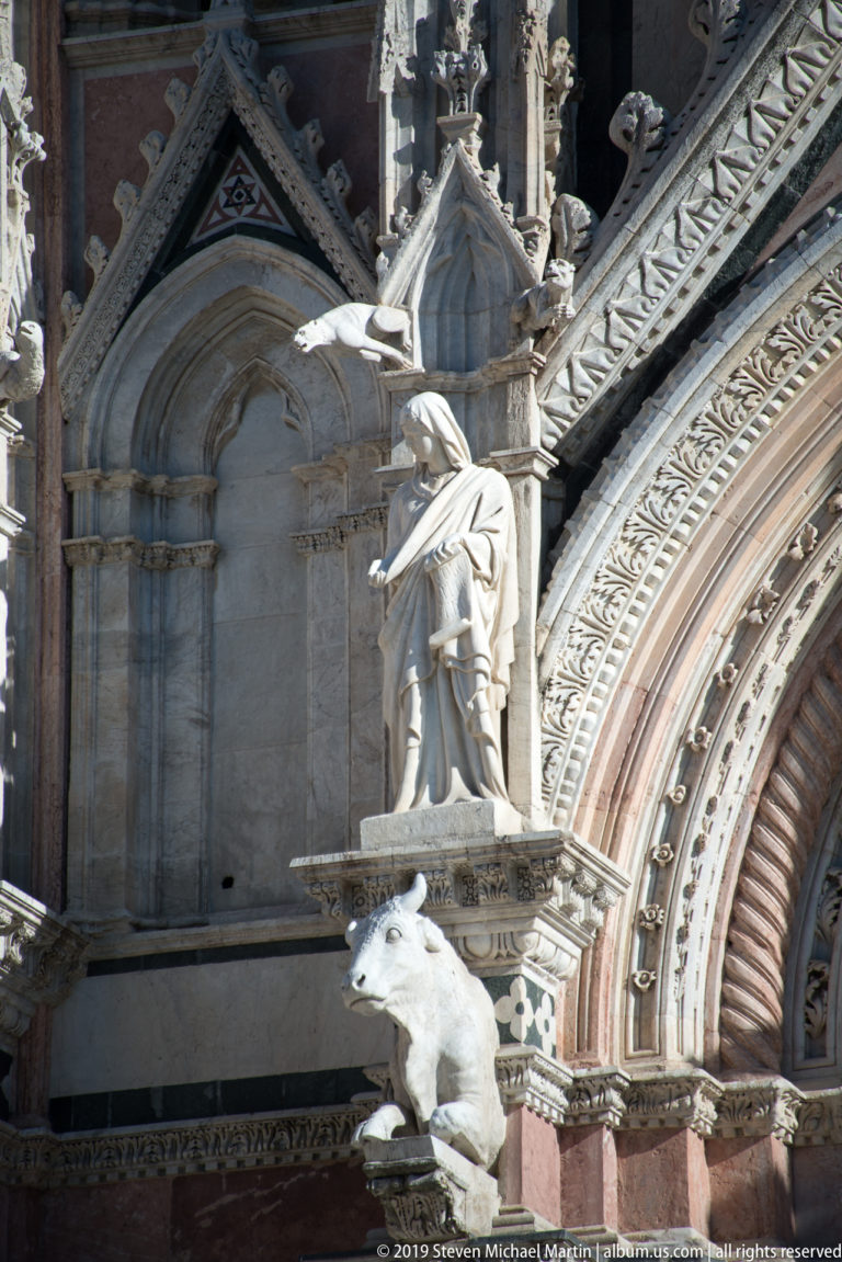 A robed stone figure stands above an animal-shaped projection in a carved cathedral façade bay.
