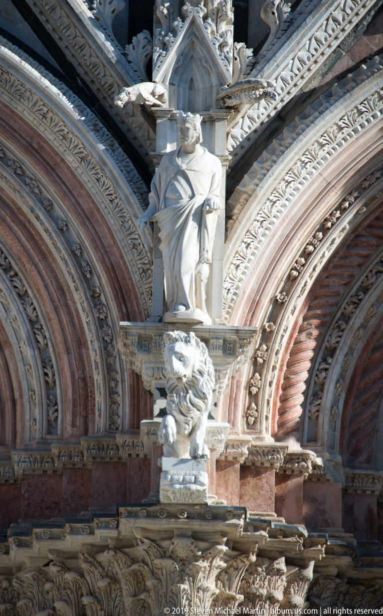 Pale carved figures project beneath a pointed canopy among striped marble and foliage carving.