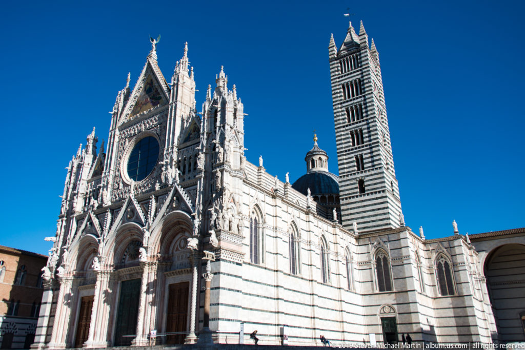 Siena Cathedral’s sculpted façade, dome, and striped campanile rise against a blue sky.