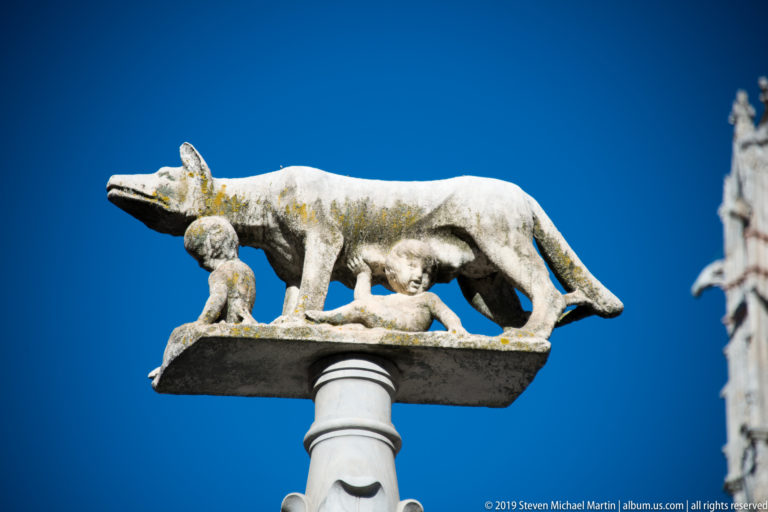 A weathered animal-like stone carving extends from a high ledge above smaller figures.