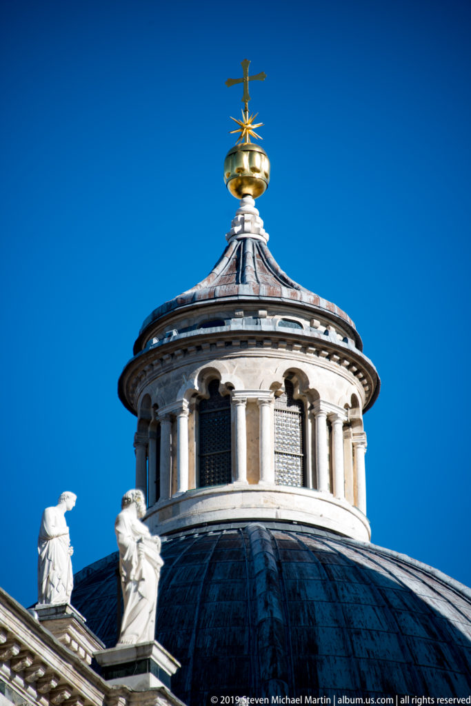 The Duomo’s dark dome and pale lantern rise to a gilded finial against a blue sky.