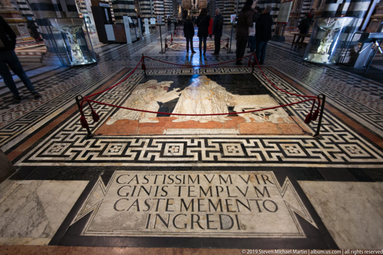 A Latin inscription in the marble floor faces visitors gathered behind a rope around an inlaid panel.