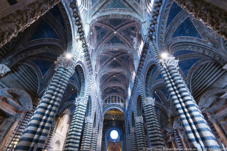 Striped columns rise into a blue starry vault that recedes toward a bright round window.