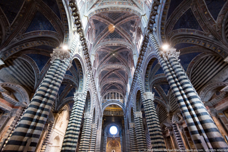 Striped piers and pointed arches rise toward a bright opening beneath the blue cathedral vault.