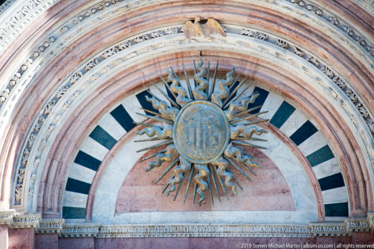 A carved human face sits within radiating dark and pale rays and blue wavy forms.