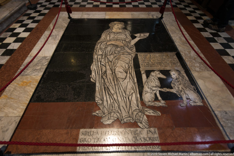 A marble inlay of a robed woman and bird stands above an inscribed plaque on the cathedral floor.
