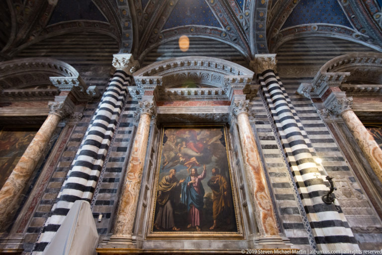 A framed religious painting and altar stand between striped marble columns beneath a blue vault.