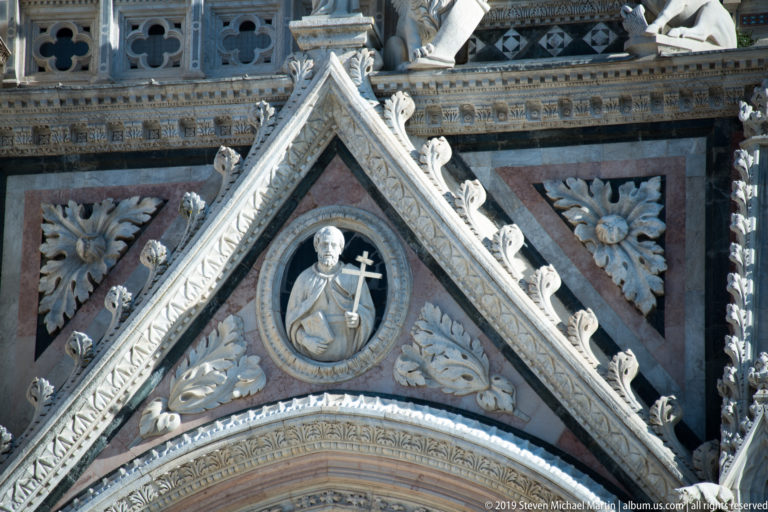 A robed stone figure holding a cross stands inside a carved cathedral niche.