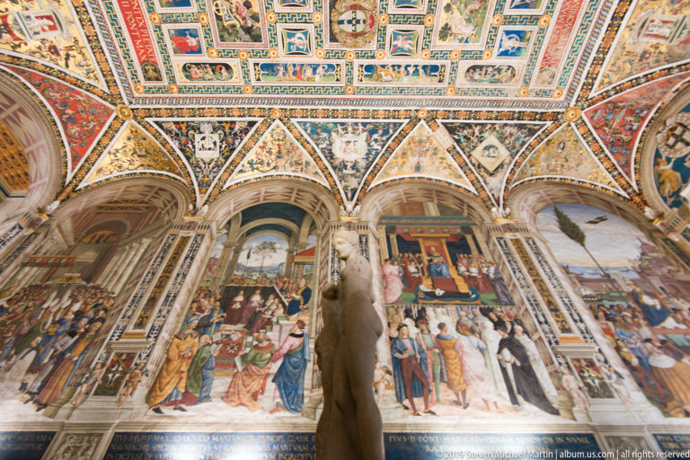 A stone figure stands before painted vault panels and arches inside Siena Cathedral.