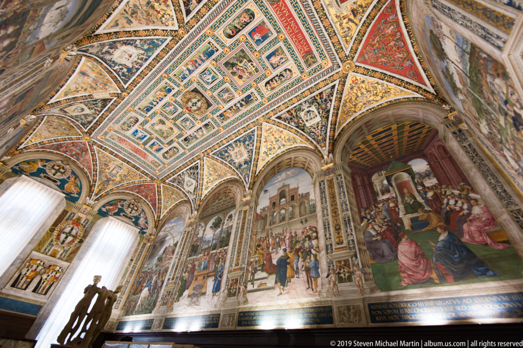 A high view across painted cathedral vaults toward patterned marble pavement and visitors below.