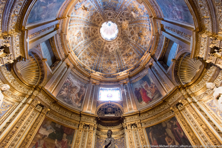 A bright circular opening surrounded by gold ornament and painted scenes on a cathedral ceiling.