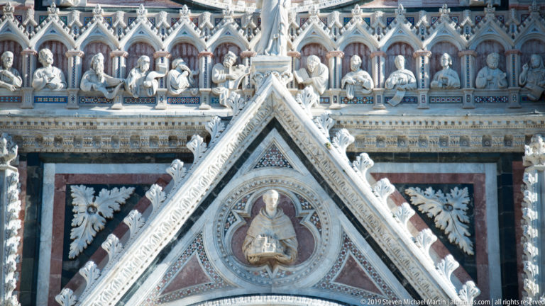 A seated robed figure holds a small model within a carved niche beneath a row of busts.